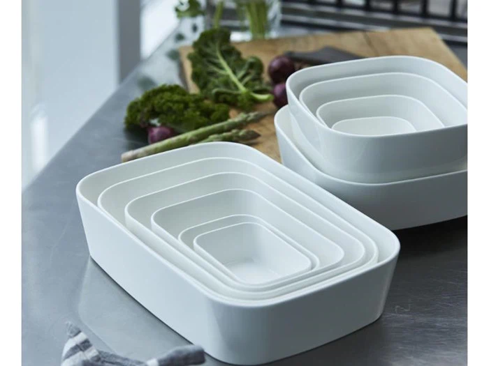 To stabler af hvide rektangulære redefade står på et køkkenbord med friske grøntsager og et skærebræt i baggrunden.