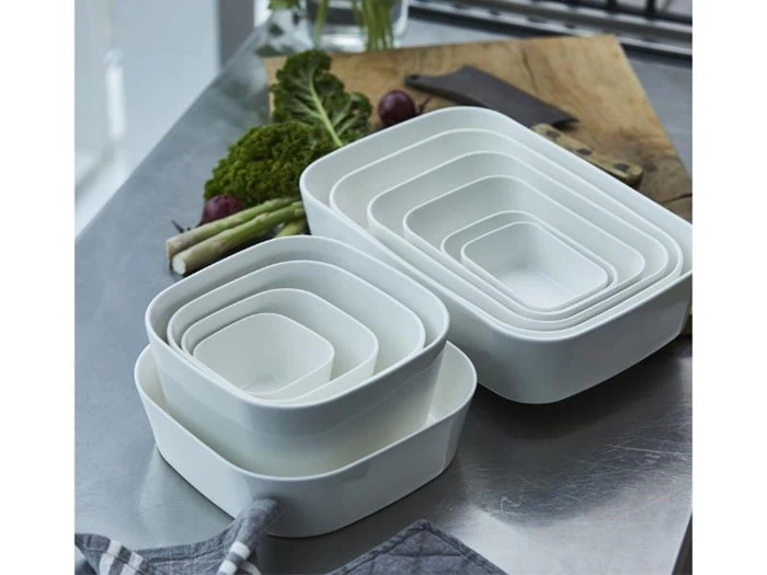 Hvide indlejrede keramiske bageforme i forskellige størrelser er stablet på et køkkenbord i rustfrit stål ved siden af friske grøntsager og et skærebræt.