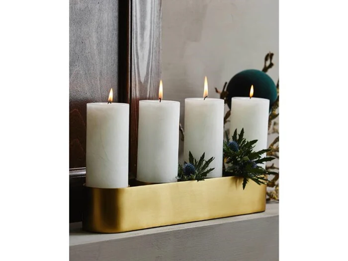 Fire hvide bloklys, der brænder i en rektangulær guldstage, dekoreret med grønt, placeret på en hvid flade nær en træramme.