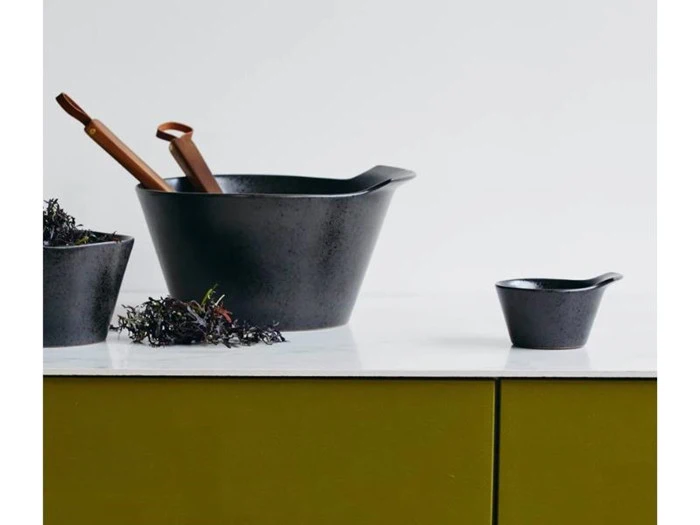 To sorte skåle står på en hvid bordplade; den ene indeholder træredskaber og tang, med grønne skabe under og en almindelig hvid baggrund.