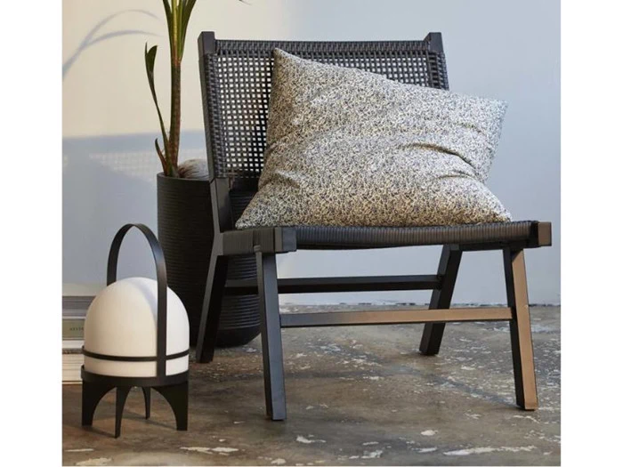 En moderne sort vævet stol med en spættet hynde står ved siden af en potteplante og en hvid, kuppelformet lanterne på et betongulv.