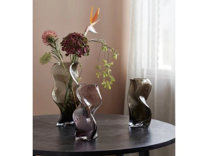 Tre bølgede, mørke glasvaser står på et rundt, sort bord; den ene vase indeholder forskellige blomster, og et blødt lys trænger ind gennem et vindue i baggrunden.