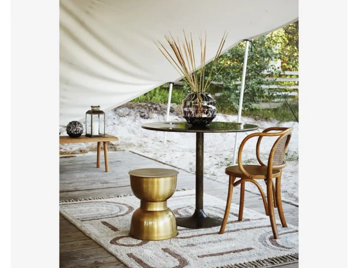 Et lille rundt bord med en dekorativ vase, en gylden skammel, en træstol og et mønstret tæppe på en træterrasse under en hvid baldakin. I baggrunden ses udendørs grønne områder.
