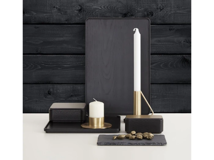 Et moderne arrangement af sorte bakker, et hvidt bloklys i en guldholder, et hvidt bloklys på en guldplade og dekorative guldgenstande på en mørk træbaggrund.