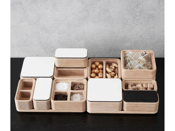 Et sæt træ- og hvide beholdere med forskellige krydderier, teposer og snacks står pænt arrangeret på en sort overflade mod en grå væg.