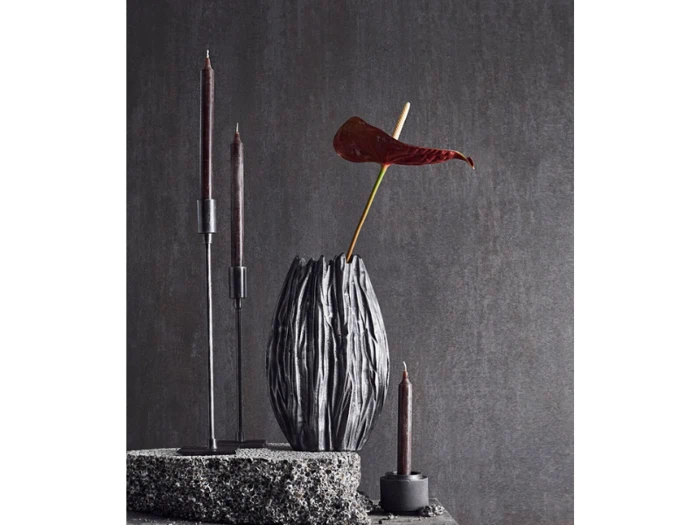 Et moderne, minimalistisk stilleben med sorte stearinlys i stager og en mørk, struktureret vase med en enkelt rød anthuriumblomst på en grå, struktureret baggrund.