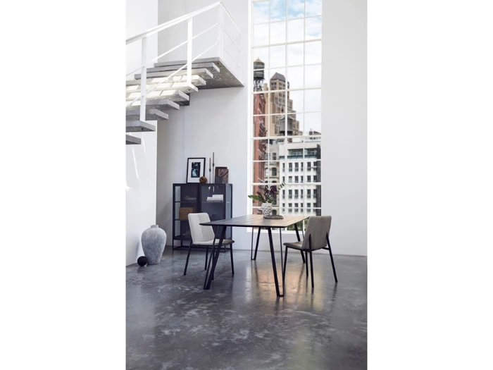 Moderne spiseplads med to stole og et bord på et betongulv, nær et stort vindue og en trappe, med byens bygninger synlige udenfor. Minimalistisk indretning med planter og kunst.