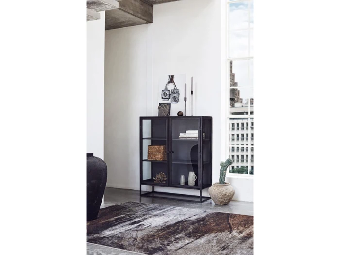 En moderne bogreol i sort metal og glas med dekorationsgenstande står ved et stort vindue i et lyst, minimalistisk rum med udsigt over byen.