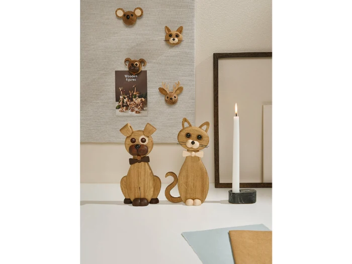 Hunde- og kattefigurer af træ med butterfly står på en hvid flade ved siden af et tændt stearinlys. Dyreformede vægdekorationer og et indrammet billede er i baggrunden.