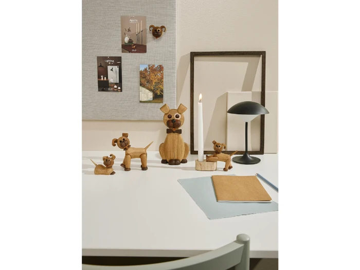 Et hvidt skrivebord med hundefigurer i træ, en sort lampe, et tændt stearinlys, en notesbog og postkort på en grå opslagstavle i baggrunden.