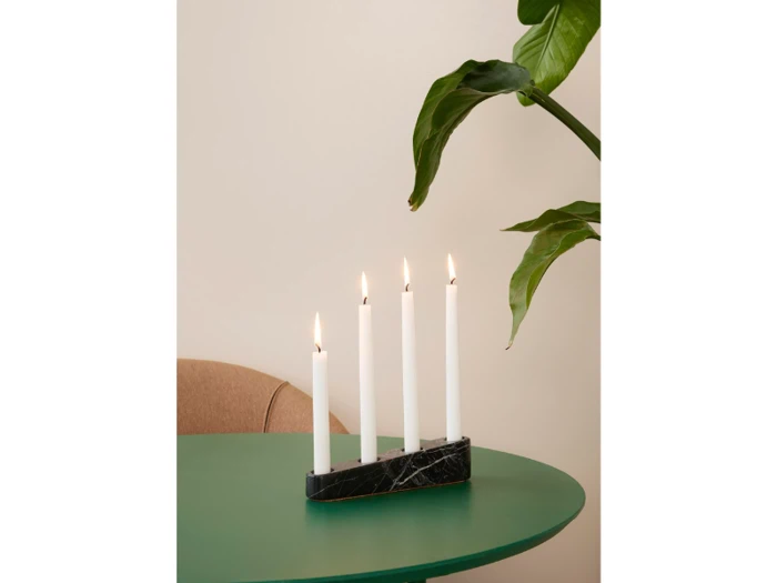 Fire tændte hvide stearinlys i en sort marmorholder står på et grønt bord med en bladplante og en del af en beige stol i baggrunden.