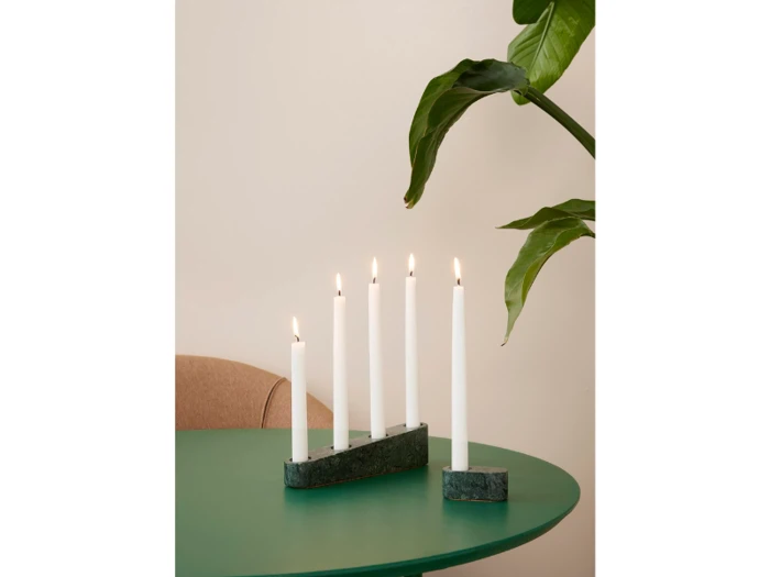 Fem tændte hvide stearinlys i marmorholdere er placeret på et grønt bord ved siden af en stor bladplante og en beige stol mod en lys væg.