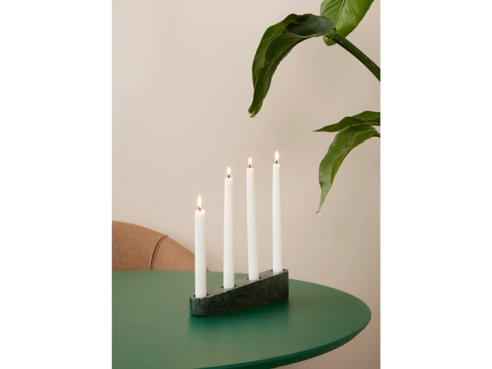 Fire tændte hvide stearinlys i en moderne grøn holder står på et rundt grønt bord med en bladplante og en del af en beige stol i baggrunden.
