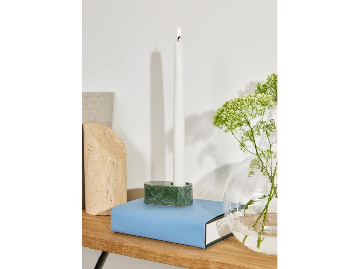 Et hvidt fyrfadslys i en grøn holder står på en blå bog på en træplade, og i nærheden står en rund vase med grønne blomster.