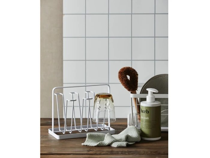 En bordplade med et hvidt opvaskestativ med to omvendte glas, en opvaskebørste, en flaske urteopvaskemiddel, en klud og tallerkener mod en flisebelagt væg.