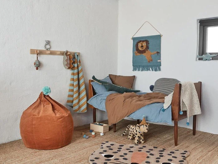 Hyggeligt børneværelse med træseng, blåt og brunt sengetøj, bamser, orange sækkestol, stribet tæppe, løvetæppe på væggen og legetøj på et vævet tæppe.