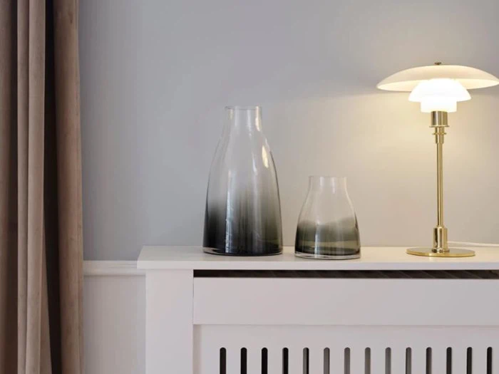 To glasvaser med gradient og en messinglampe med hvid skærm står på en hvid radiatorskjuler mod en lysegrå væg med beige gardiner til venstre.