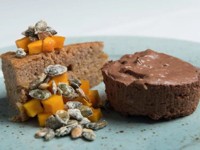 En tallerken med et stykke kage toppet med mango i tern og kerner ved siden af en rund portion chokolademousse.