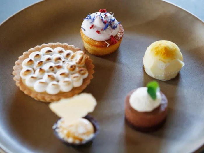 En brun tallerken med fem forskellige desserter i mundrette størrelser, bl.a. en tærte med ristet marengs, en mini-cupcake med creme og en rund hvid trøffel.