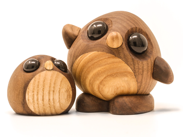 To runde uglefigurer af træ med store sorte øjne og udskårne næb, den ene lidt større og oprejst, den anden mindre og liggende, på en hvid baggrund.