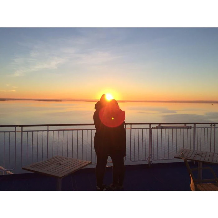 To mennesker står tæt sammen på en terrasse ved solopgang eller solnedgang og vender ud mod et roligt vandområde med solen i horisonten, der skaber en varm glød omkring dem.