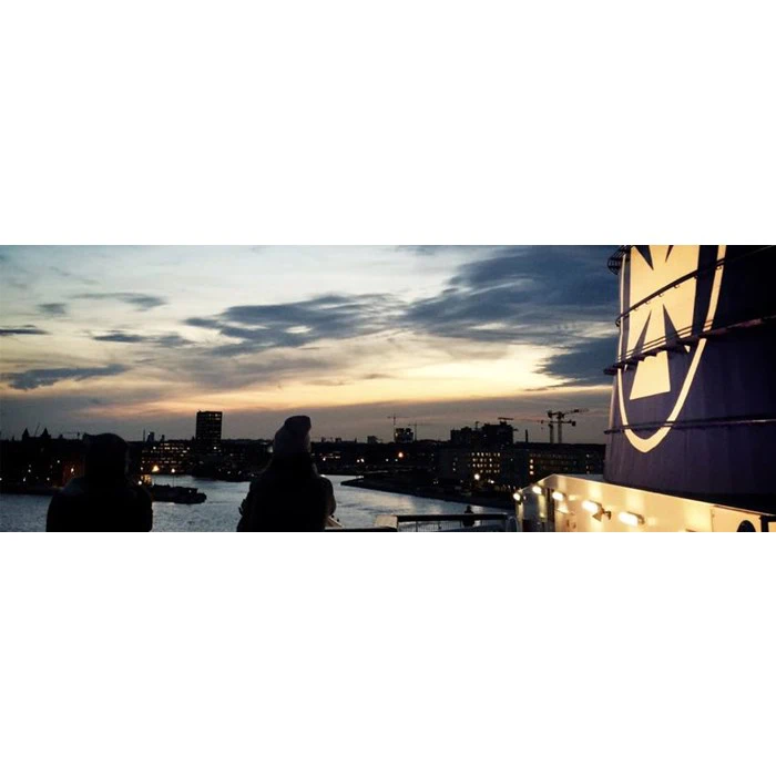 To mennesker står på et skibsdæk ved solnedgang med udsigt til en bys skyline med bygninger og kraner under en overskyet himmel.