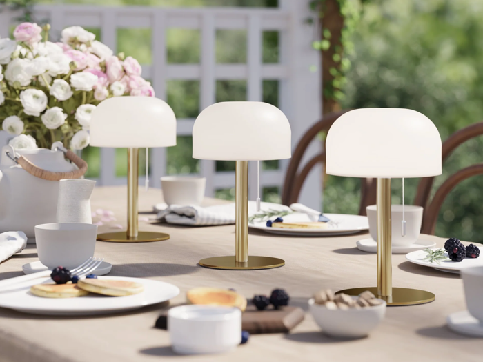 Et elegant bord dækket udendørs med tre moderne lamper i hvidt og guld, tallerkener, kopper, bær, pandekager og en blomsterbuket i baggrunden.