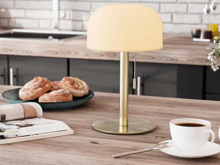 En bordlampe af messing med en rund lampeskærm står på køkkenbordet ved siden af en kop kaffe, wienerbrød på en tallerken og et åbent blad.