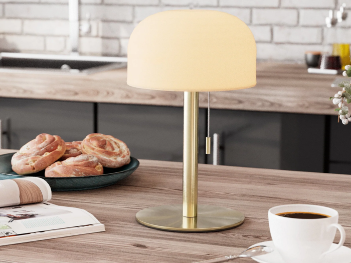 En bordlampe af messing med en hvid lampeskærm står på en køkkenbordplade af træ ved siden af kager, en kop kaffe og et åbent magasin.