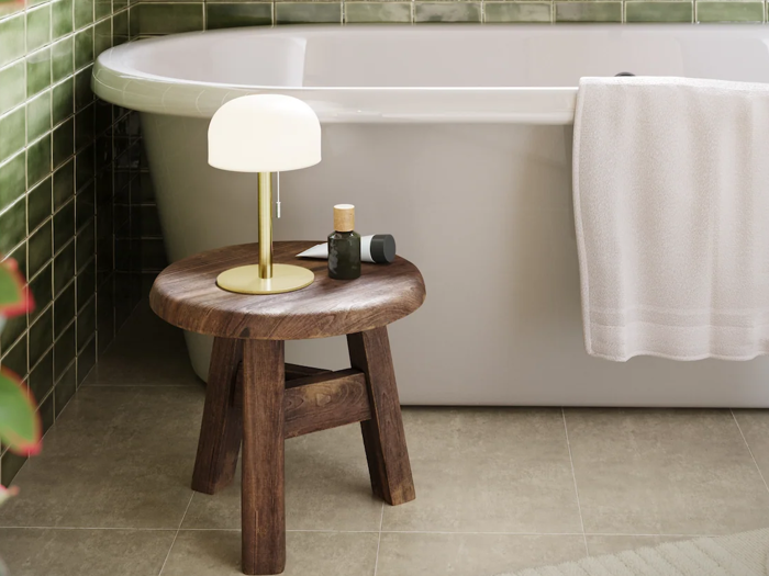 Ved siden af et hvidt badekar, hvor der hænger et håndklæde over kanten, står en træskammel med en lampe og toiletartikler; væggene er beklædt med grønne fliser, og gulvet er belagt med beige fliser.