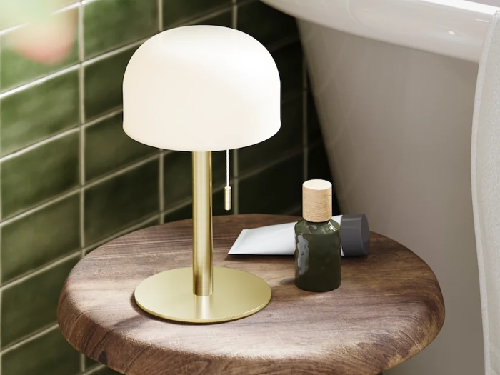 En lille guldlampe med en hvid lampeskærm står på et rundt træbord ved siden af en grøn flaske og et hvidt rør, foran en væg med grønne fliser og et hvidt badekar.
