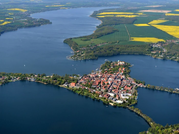 Luftfoto af en lille by på en halvø ved søen, omgivet af vand og grønne marker med gule pletter under en klar himmel.