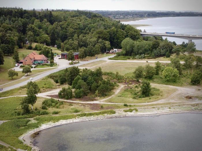 Luftfoto af et område ved en sø med snoede stier, grønne træer, flere bygninger og en bro i det fjerne under en overskyet himmel.