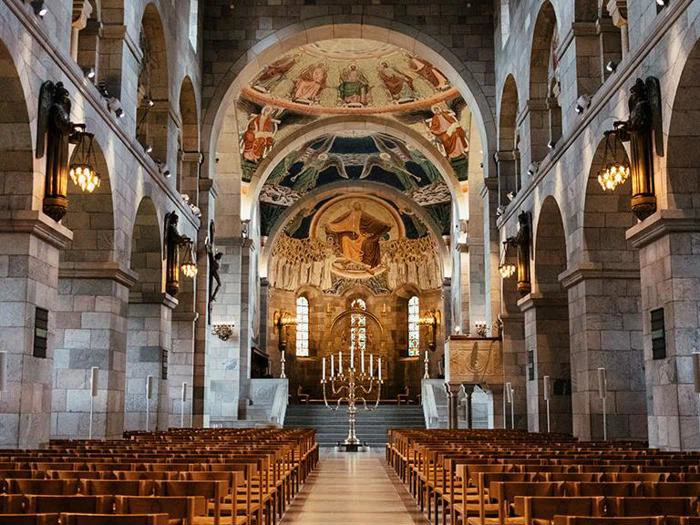 Interiør i en stenkirke med rækker af træbænke, udsmykkede lysekroner, buegange og farverige religiøse vægmalerier over alteret.