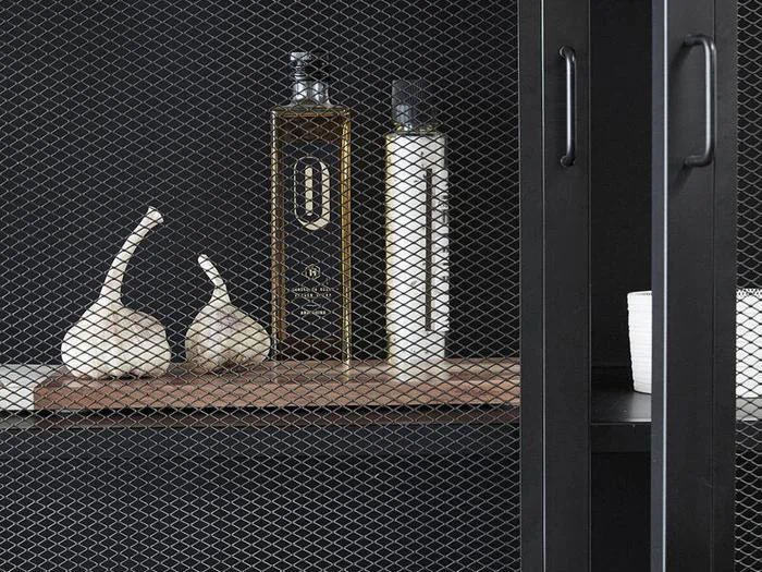 To hvidløgsløg, en flaske olivenolie og en anden høj flaske står på en hylde bag en skabsdør af sort metalnet.