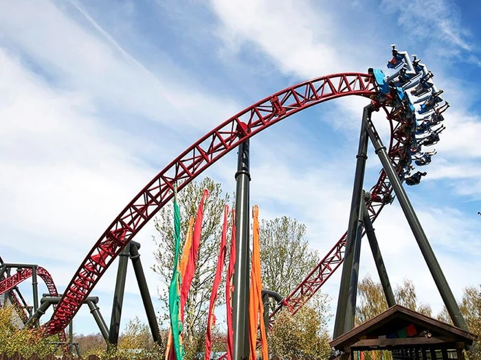 En rutsjebane i rødt stål looper højt oppe i himlen med passagerer, der kører, omgivet af træer og farverige serpentiner nedenfor under en delvist overskyet himmel.