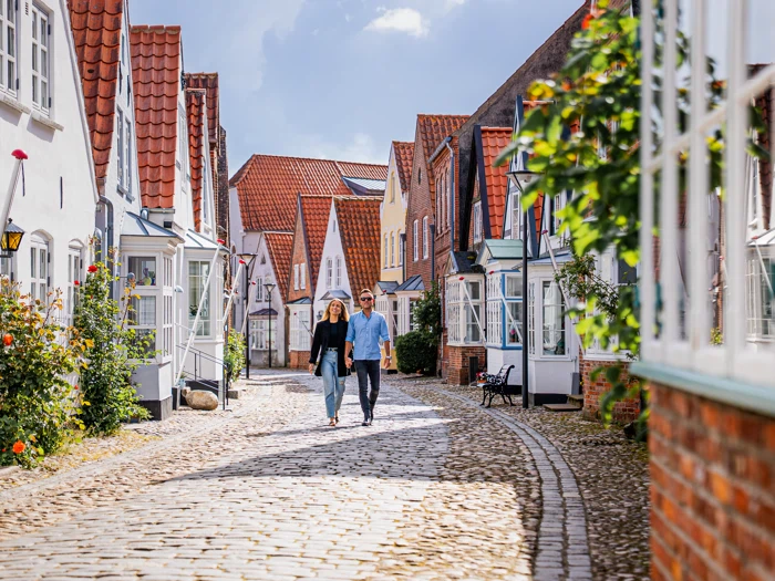 Et par går ned ad en brostensbelagt gade med charmerende huse med røde tegltage og hvide facader på en solrig dag.