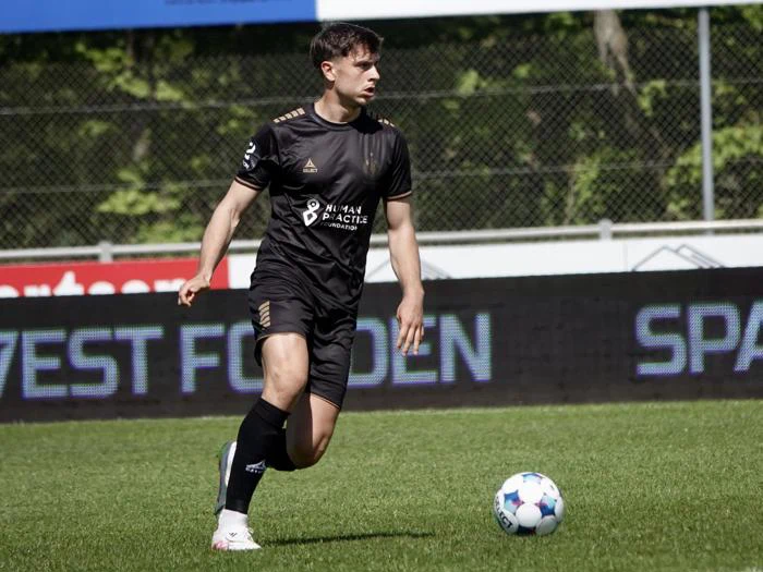 En mandlig fodboldspiller i sort uniform løber på en græsbane, mens han styrer en blå og hvid fodbold. Et indhegnet område og træer er synlige i baggrunden.