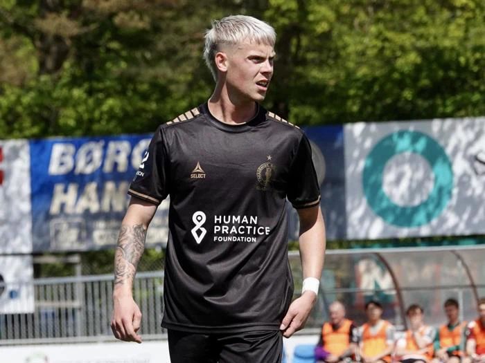 En fodboldspiller med lyst hår og tatoveringer bærer en sort trøje og står på en bane under en kamp med træer og tilskuere i baggrunden.