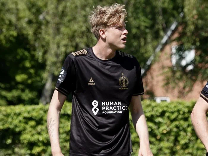 En fodboldspiller i en sort trøje står udendørs på en solskinsdag og kigger til siden med grønne områder og en bygning i baggrunden.