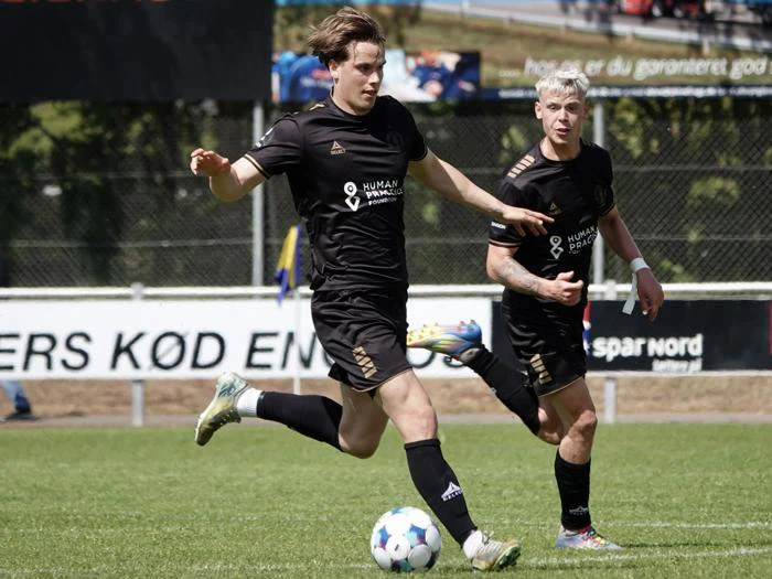 To fodboldspillere i sorte uniformer står på en græsbane under en kamp. Den ene sparker til en blå og hvid bold, mens den anden løber ved siden af ham. I baggrunden ses et hegn og bannere.