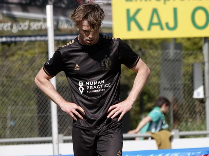 En fodboldspiller i en sort trøje står på banen med hænderne på hofterne, kigger ned og ser træt eller skuffet ud. Et gult skilt og en tilskuer er synlige i baggrunden.