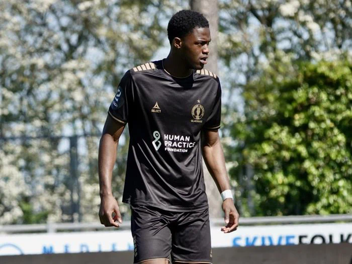 En fodboldspiller i en sort og guld uniform står på en bane, fokuseret og klar, med træer og et sløret skilt i baggrunden.