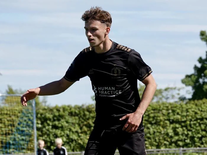 En mandlig fodboldspiller i sort uniform står på en græsbane og forbereder sig på at bevæge sig. Træer og en målstolpe er synlige i baggrunden.