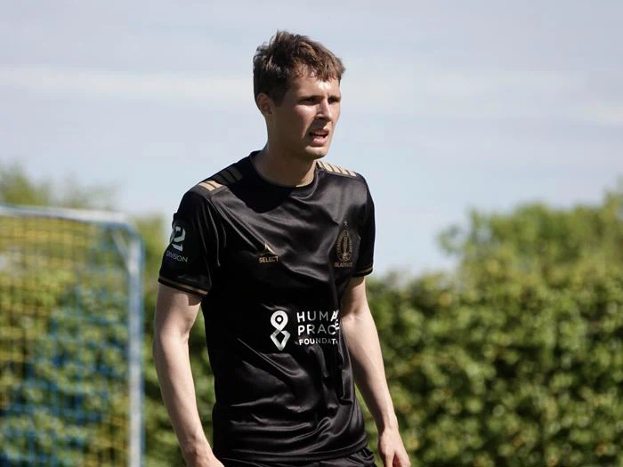 En mandlig fodboldspiller i sort trøje står på en bane med grønne områder og en målstolpe i baggrunden og ser fokuseret ud.