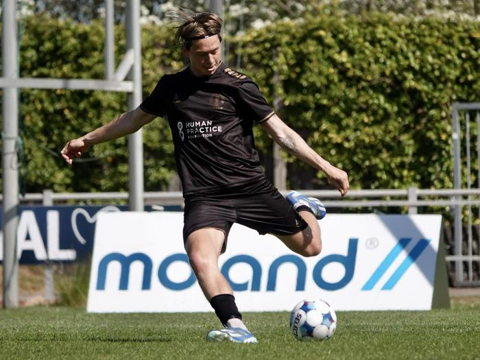 En fodboldspiller i sort uniform sparker til en fodbold på en græsplæne med grønne områder og et skilt i baggrunden.