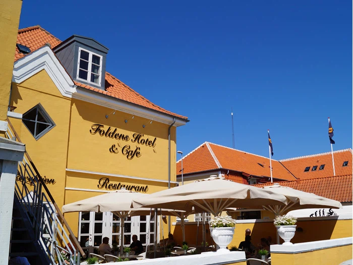 Gult hotel og café med orange tegltag, udendørsservering under store parasoller og klar blå himmel over. På skiltene står der "Folkens Hotel & Cafe" og "Restaurant".