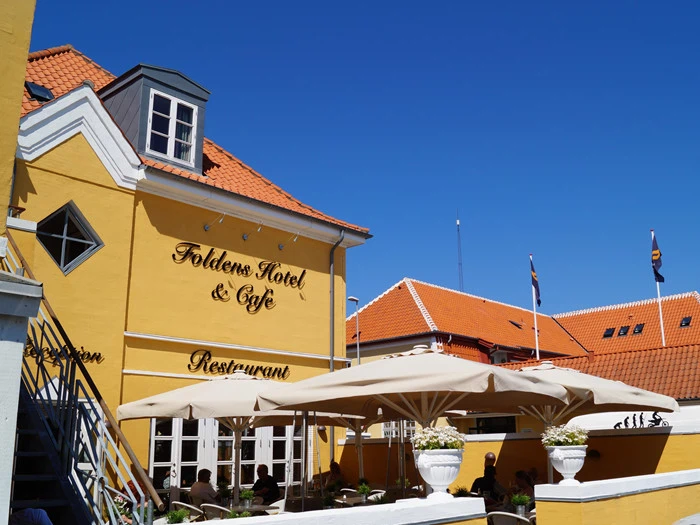 Gul bygning med Foldens Hotel & Café Restaurant på facaden, røde tegltage, store hvide parasoller og mennesker, der spiser udenfor under en klar blå himmel.