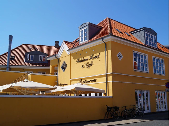 Et lysegult hotel og café med rødt tegltag, udendørs siddepladser under parasoller og cykler parkeret foran på en solrig dag.