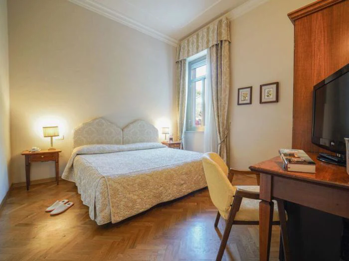 Et hyggeligt hotelværelse med en dobbeltseng, natborde med lamper, et vindue med gardiner, et skrivebord med stol, et tv og tøfler på trægulvet.